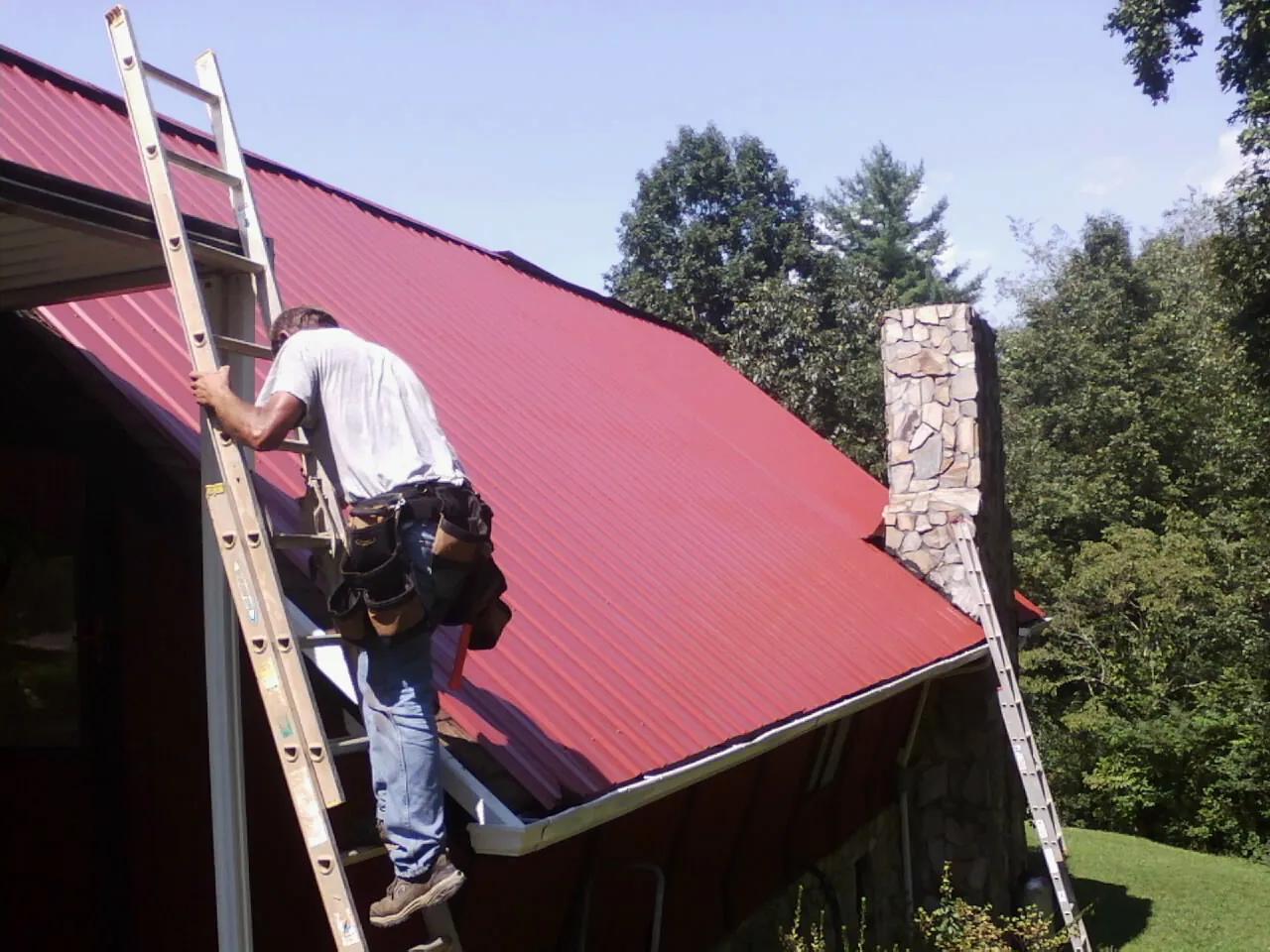 Roofer on a ladder beside a red metal roof and stone chimney — Soffit & Fascia Repair by Roofing Brothers in Mastic Beach