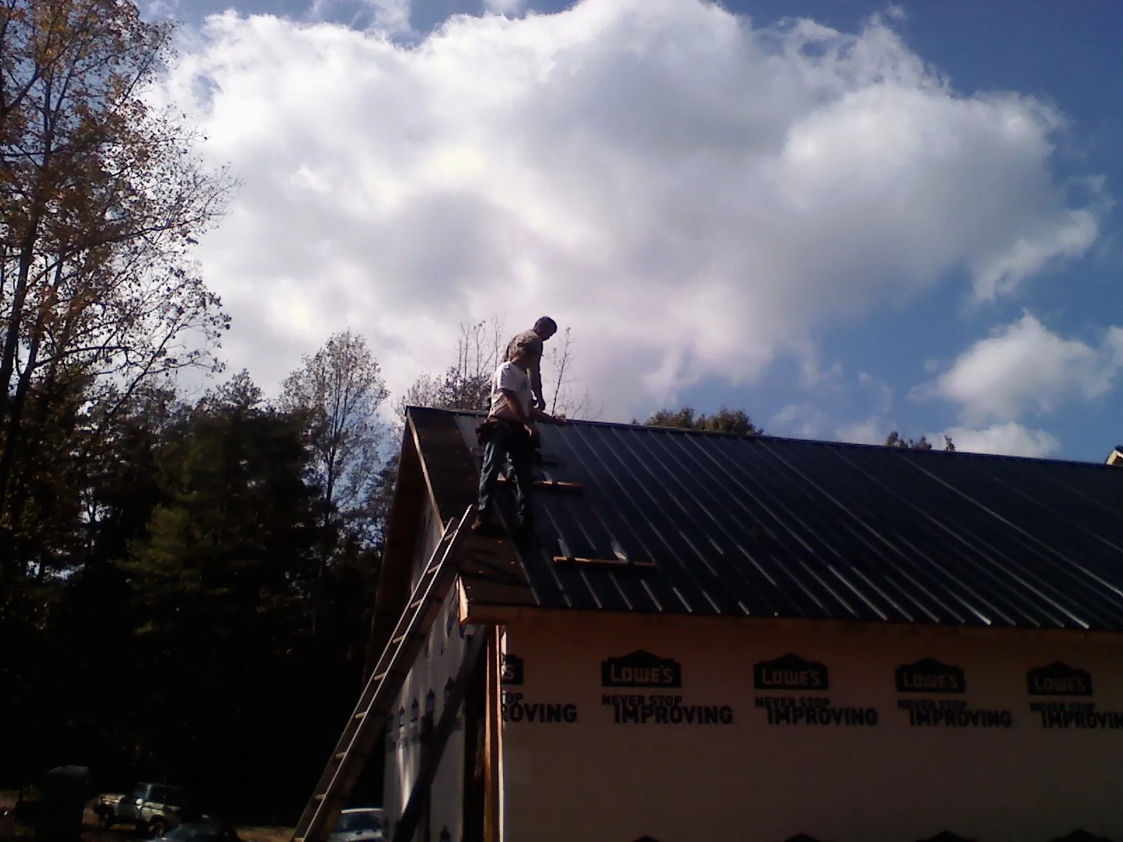 Roofer fastening dark metal panels high against a cloudy sky, from a Roof Inspection job in Mastic Beach